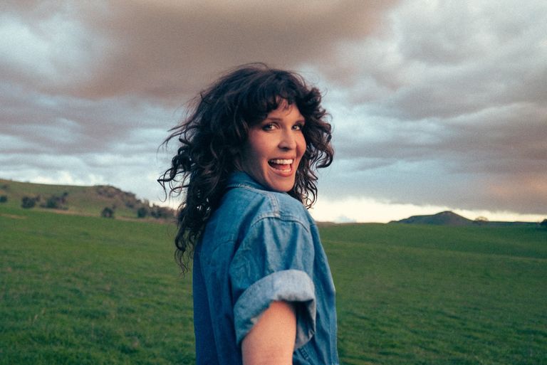 Country singer Fanny Lumsden smiling in a denim shirt, looking back over her shoulder in a green rural field beneath a cloudy sky.